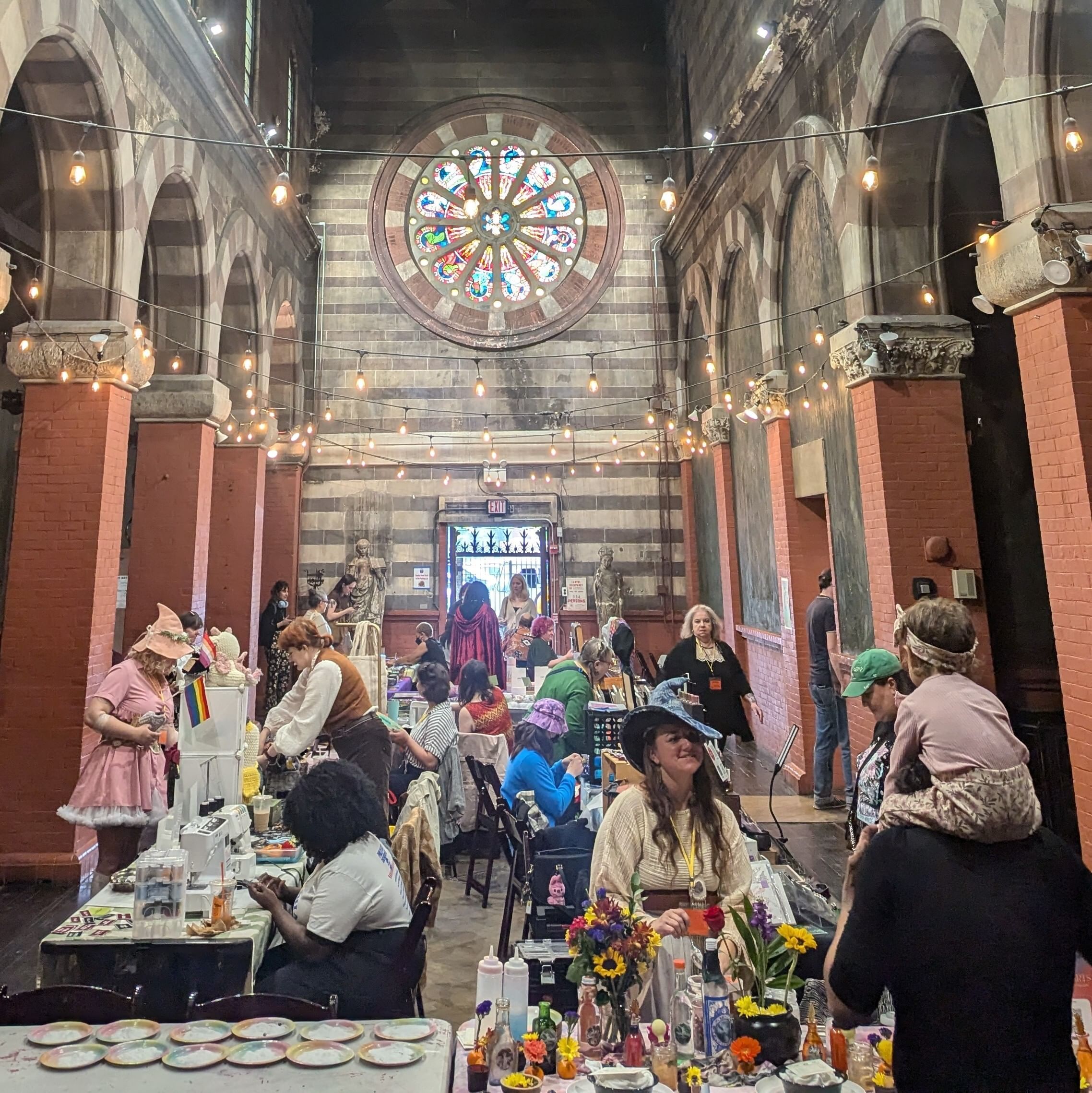 A group of vendors sit at their booths in costumes inside a building with a stained glass window.