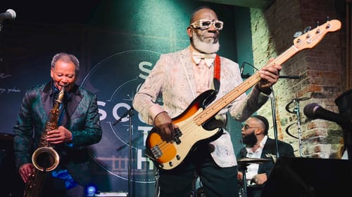 In stylish, fanciful suit jackets and bowties, the musicians play on a small stage with a brick wall to the right.