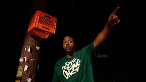 Munir, a Black man in a green tee, next to a utility pole with an orange Wawa crate on it, points in a dynamic pose.