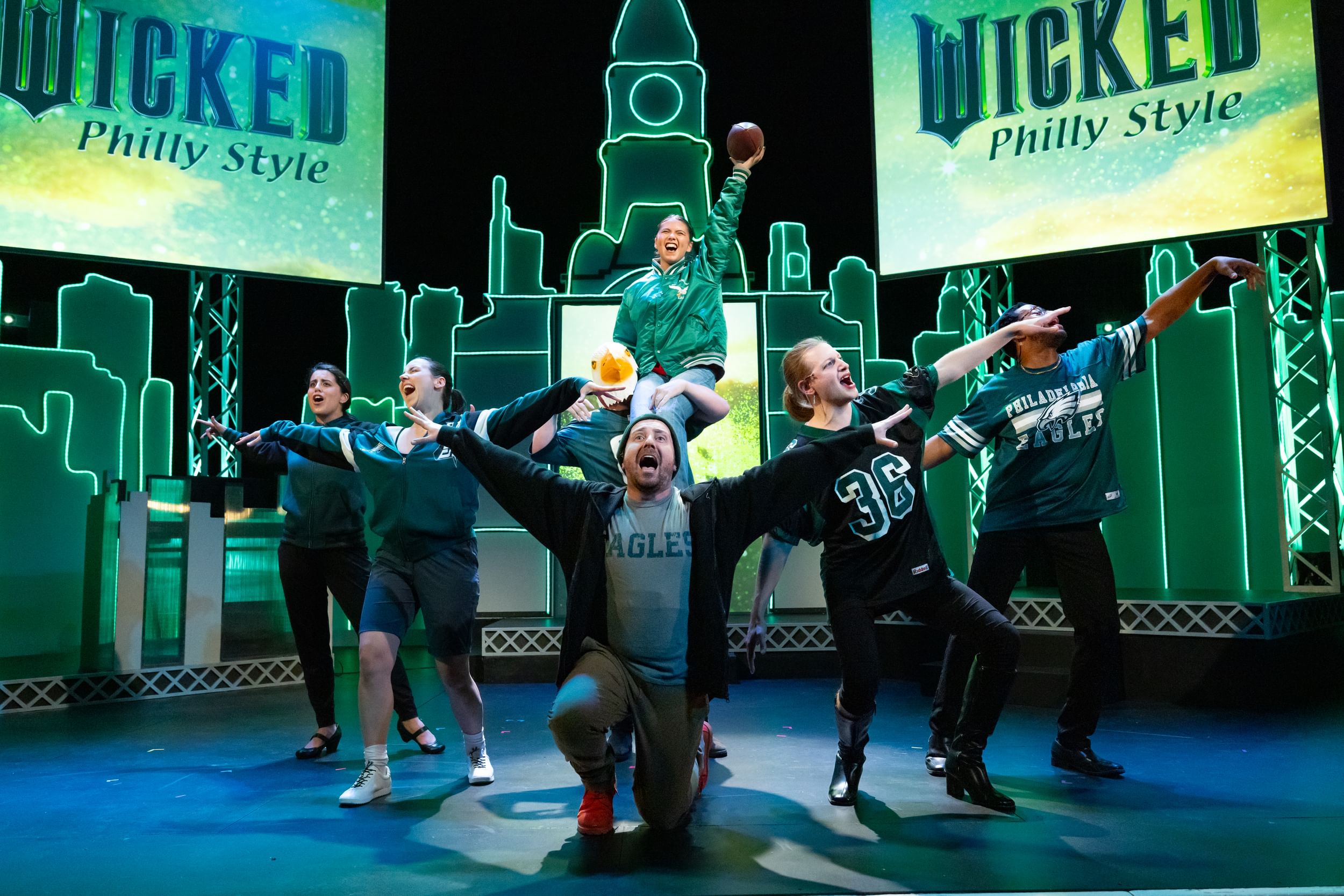 7 actors pose in a jubilant V, wearing Eagles gear, against a green neon Philly skyline & “Wicked Philly style” projections