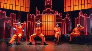 On an orange-lit set with neon bars & a Philly skyline backdrop, four actors in orange jumpsuits pose outlandishly on chairs.