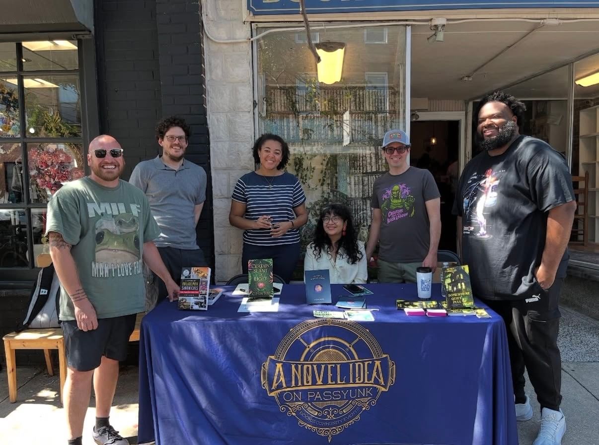 In dappled shade under a sidewalk tree, the authors pose together smiling around a table with a blue cloth.