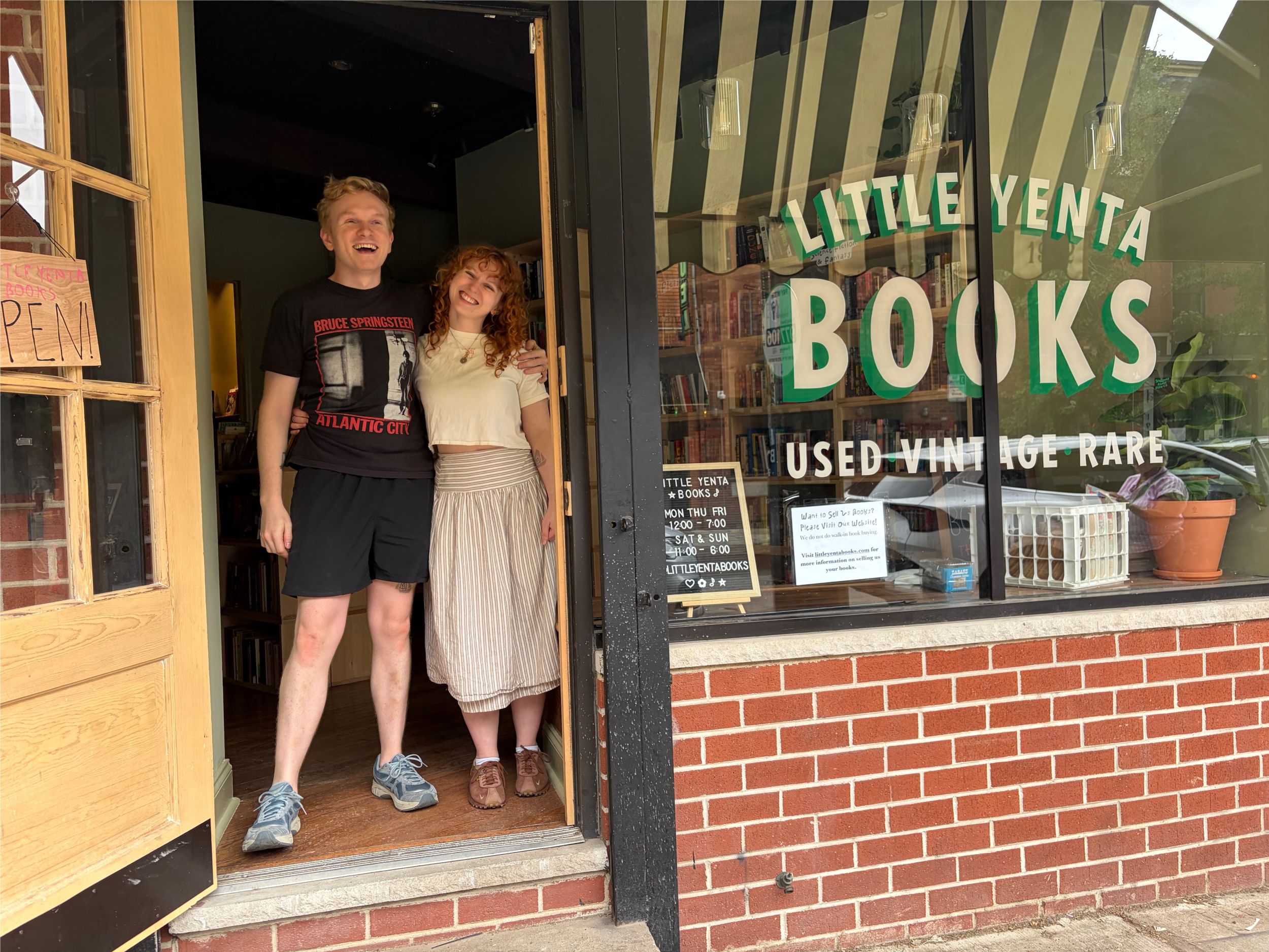 The Censors, a young white couple, pose together smiling in the door of their small storefront with green & white letters.