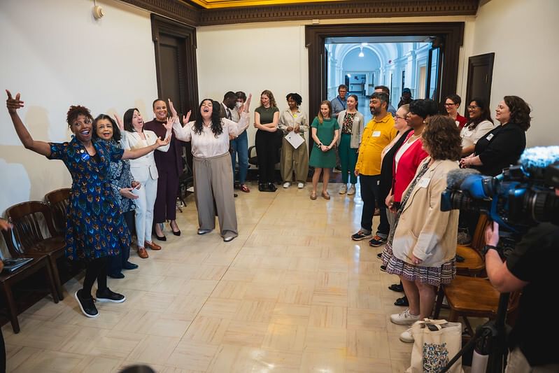 In a wide hallway at City Hall, smiling arts leaders strike a pose with arms upraised, next to a crowd of attendees.