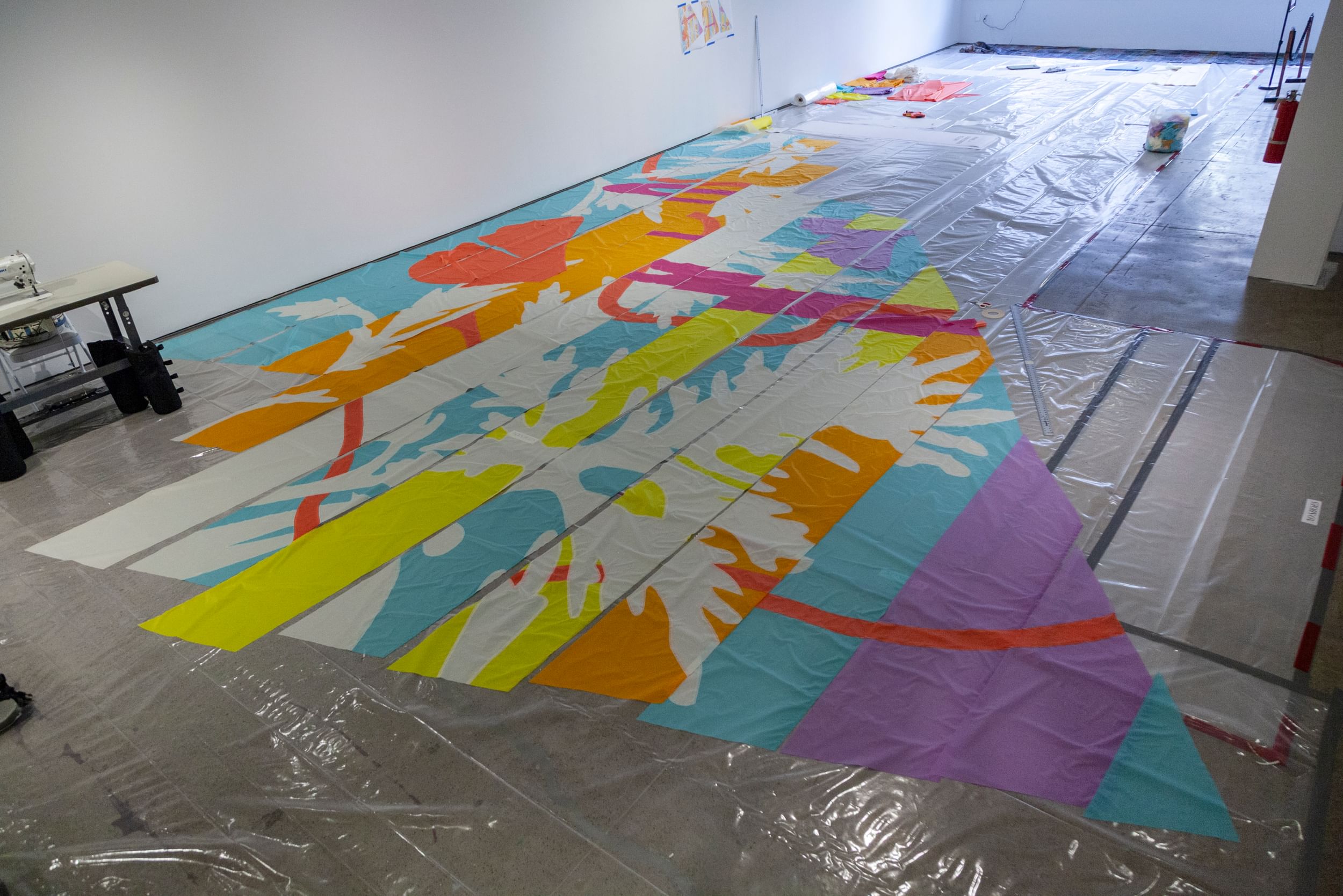 A huge triangular sail in pieces on the studio floor, with purple, blue, orange, yellow & white patterns and stripes.