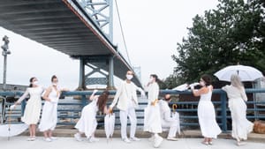 A group of dancers pose, all in white. Everyone has umbrellas, one of them is open. The Ben Franklin bridge is behind them.