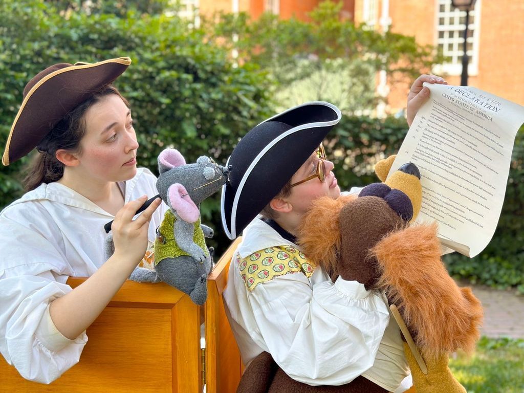 Closeup on 2 people in 18th-century garb holding puppets that interact with a copy of the Declaration outside on a sunny day