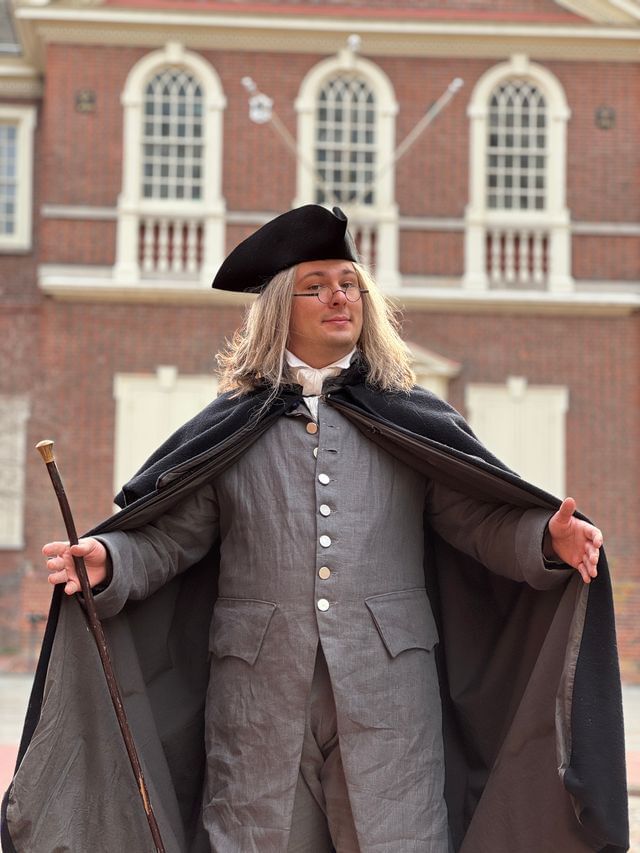 A young man with round glasses, long hair, and gray 18th-century garb stands with his arms extended in welcome.