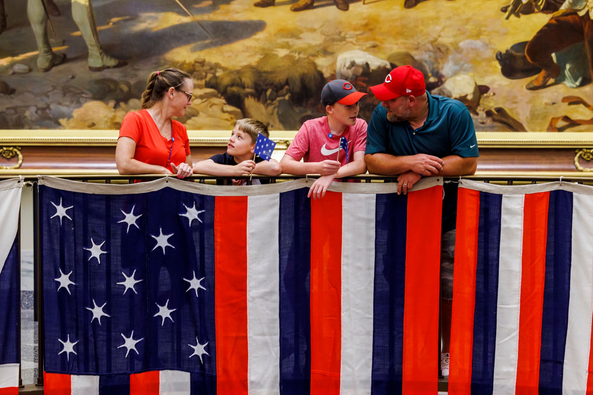 Family of two adults & two kids chat happily with each other while leaning on a balcony draped with a stylized American flag