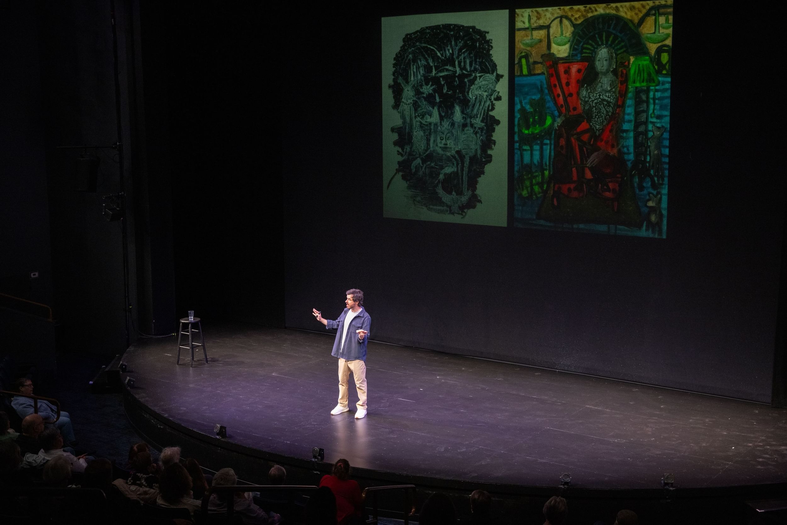 Kissajukian, in khakis & open blue button-down, stands on an empty stage with two large, surreal paintings projected above