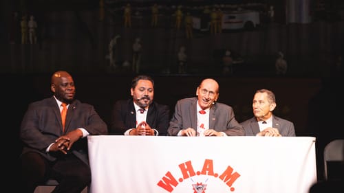 The 4 sit looking serious at a table with a cloth that says W.H.A.M.: White Hetero Affluent Men.