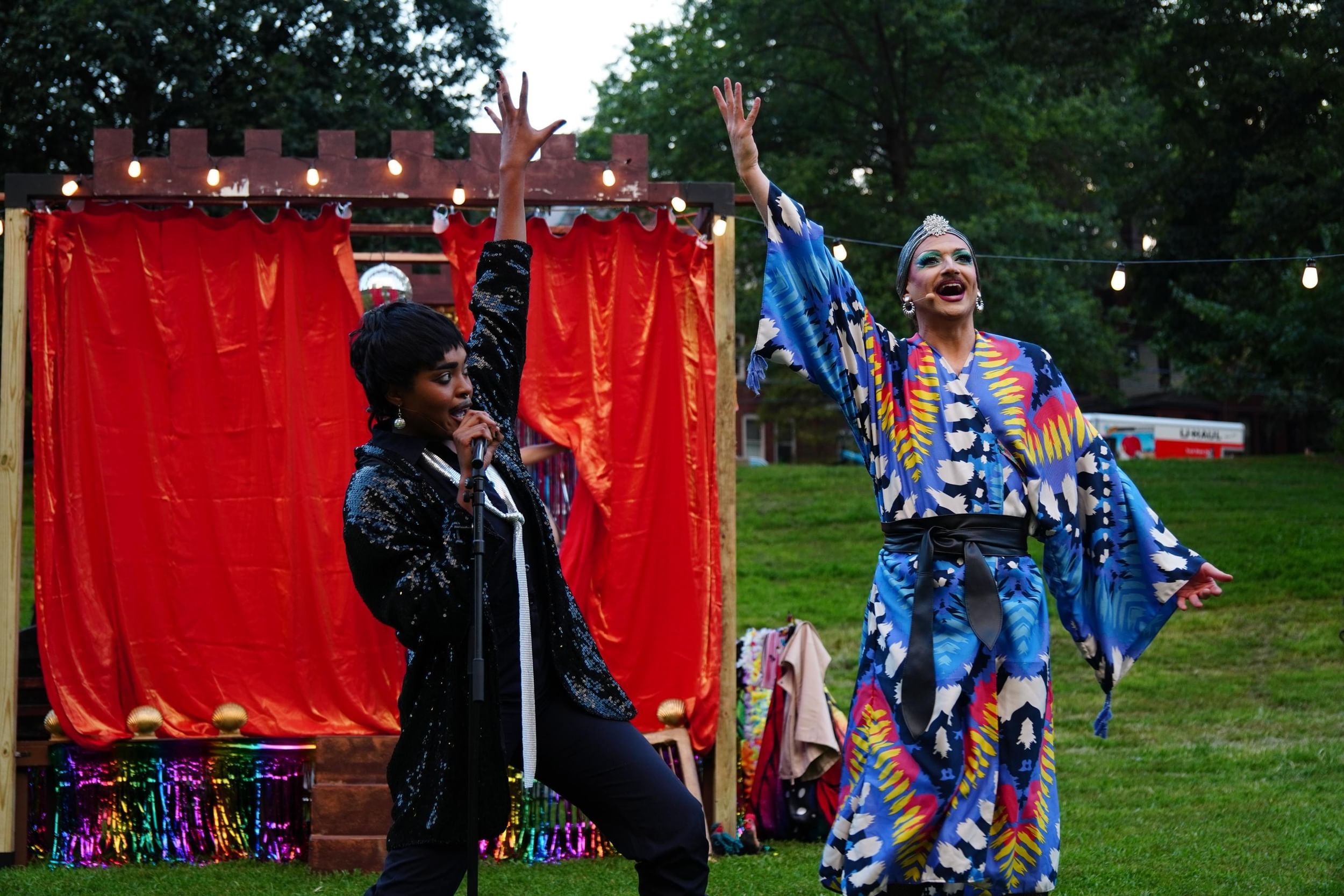 Rayne and St. Queer sing exuberantly with one hand aloft, Rayne in a sequined black blazer & St. Queer in a patterned kimono