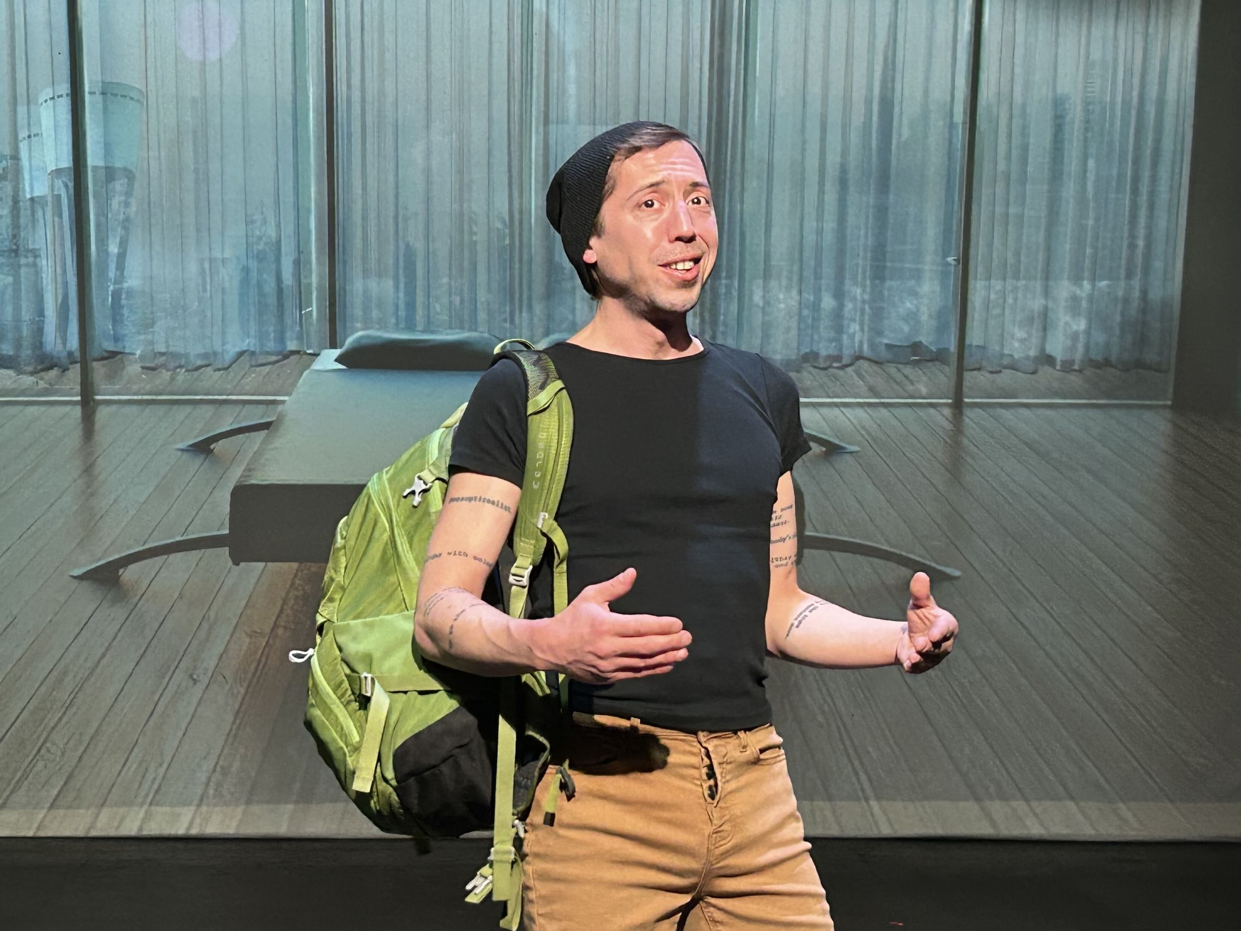 Carboni, a white man in black tee, khakis, and a green backpack, speaks to the viewer against a surreal hotel-room backdrop