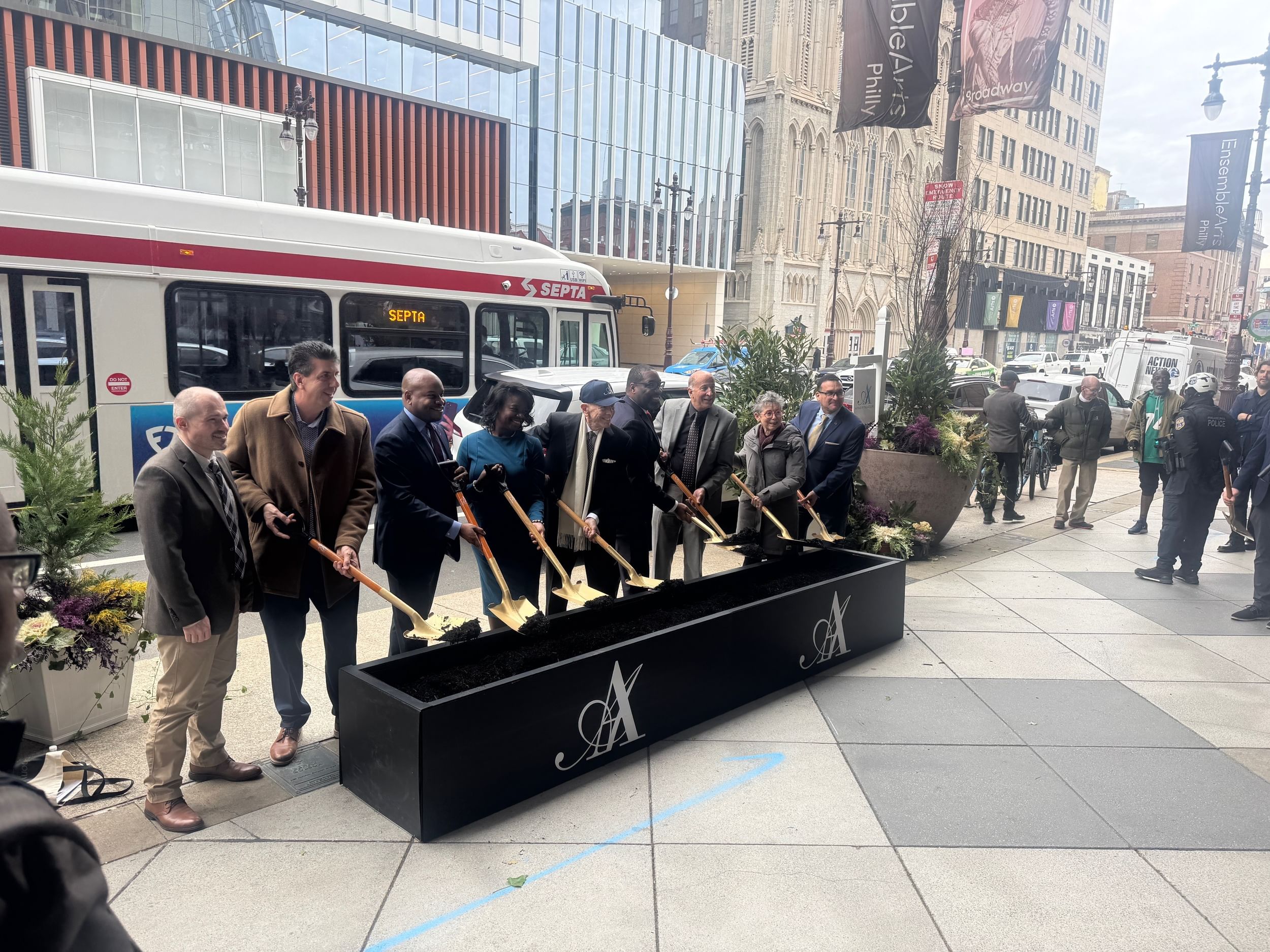 Nine people pose with dirt on golden shovels above a long planter on the city sidewalk, with a bus behind them.