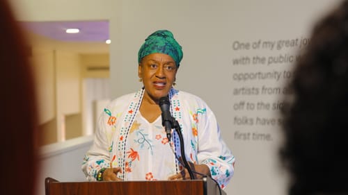 Pounder speaks at a podium in the gallery. She wears a white robe with bright floral embroidery and a turquoise turban.