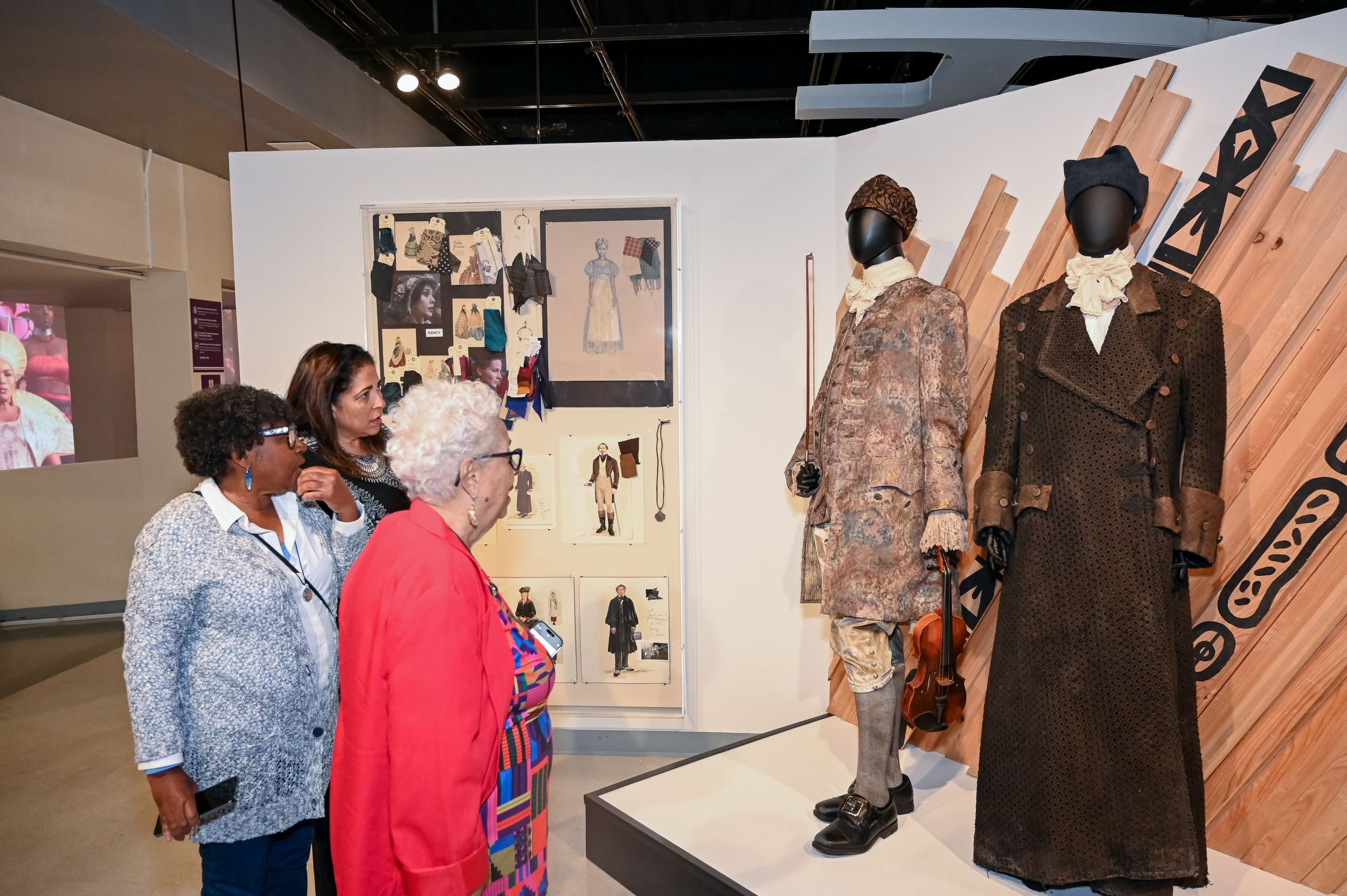Three people at left look at a display of two detailed 18th-century style men’s costumes in richly patterned earth tones.