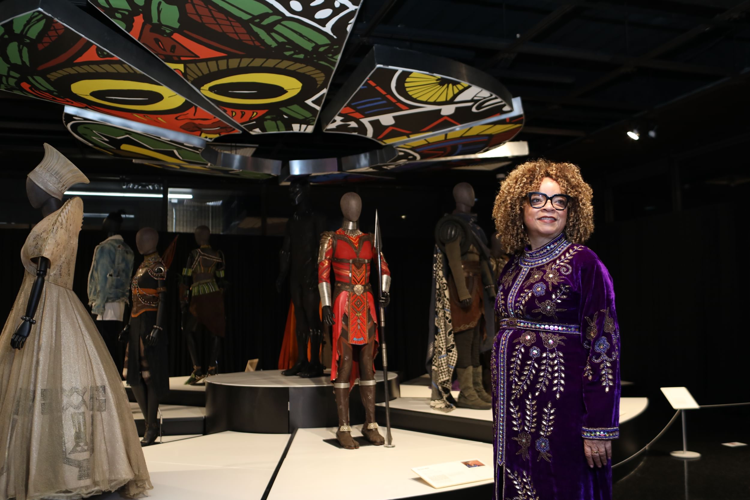 Carter, in a purple velvet robe with ornate floral embroidery, stands smiling next to a display of her Black Panther costumes