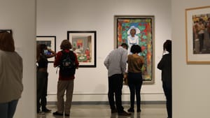 In one section of a white-walled gallery, six people with their backs to the camera examine several colorful artworks.