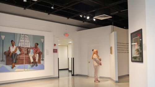 At left, a large-scale realistic painting of two people on a couch. A woman in a pale suit looks at a smaller work nearby.