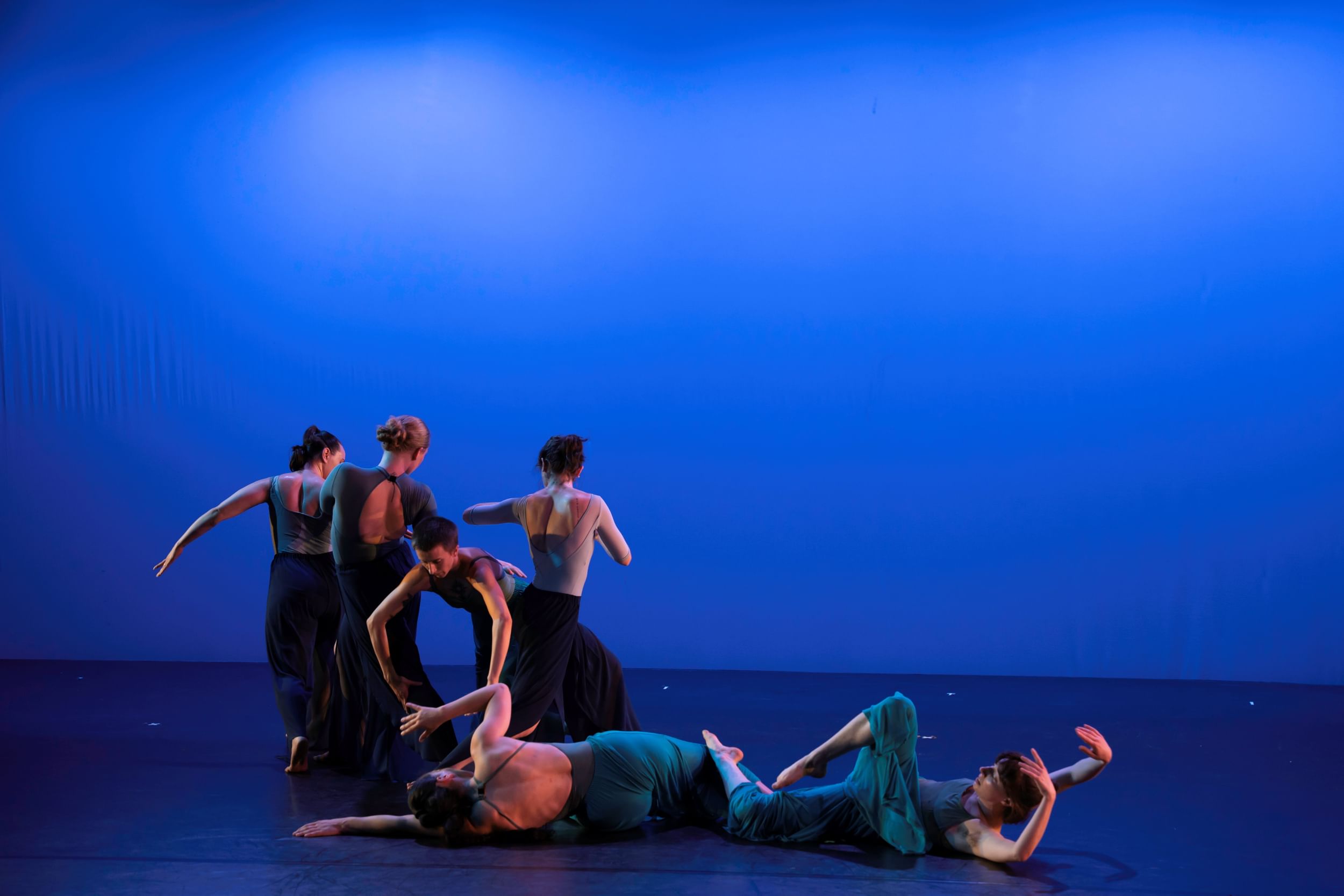 On a blue-lit stage, 4 dancers stand and 2 lie on the floor, creating a fluid sculpture with their bodies curving and limbs.