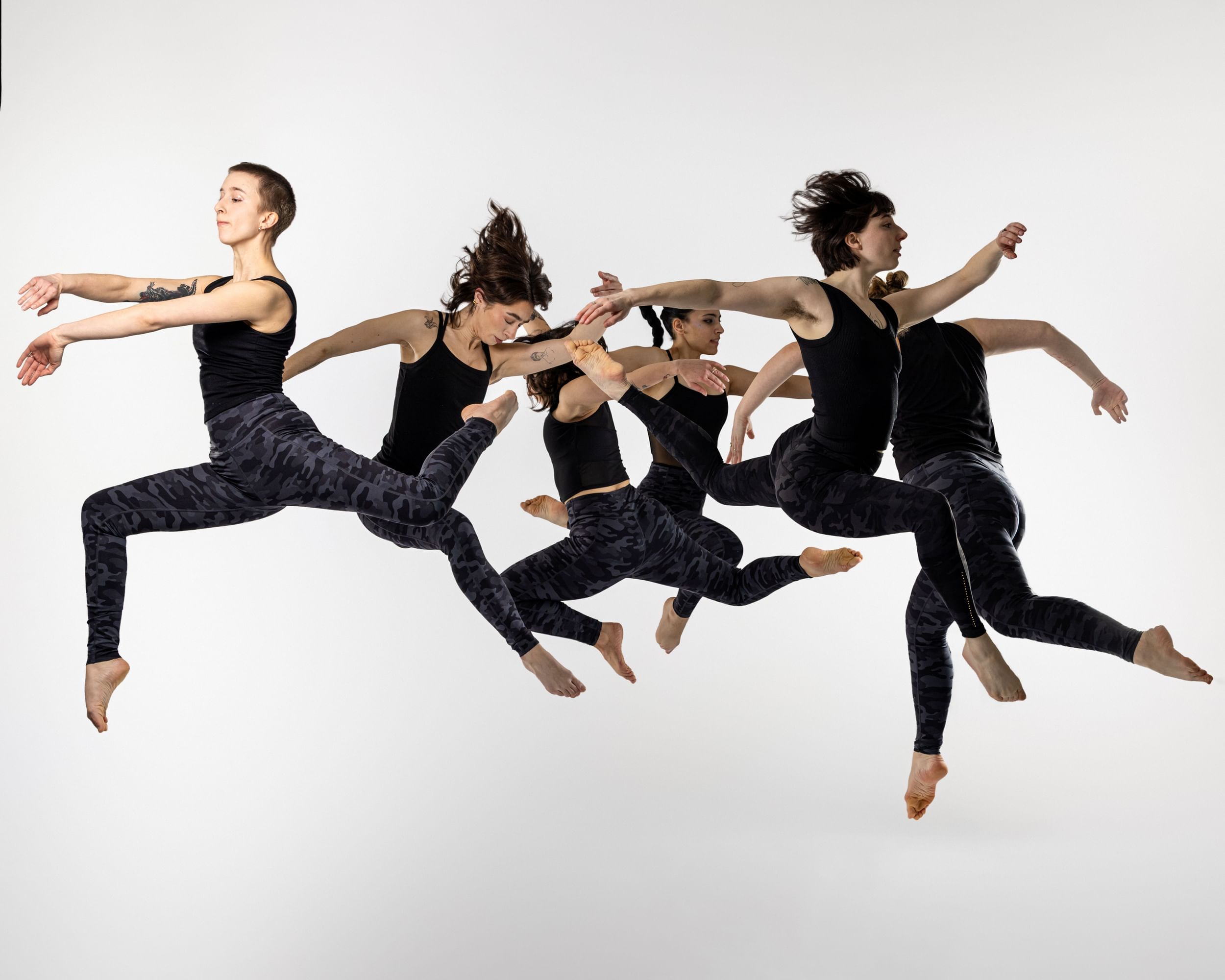 Six dancers in all black leap in the air, in various poses, against a solid white backdrop.