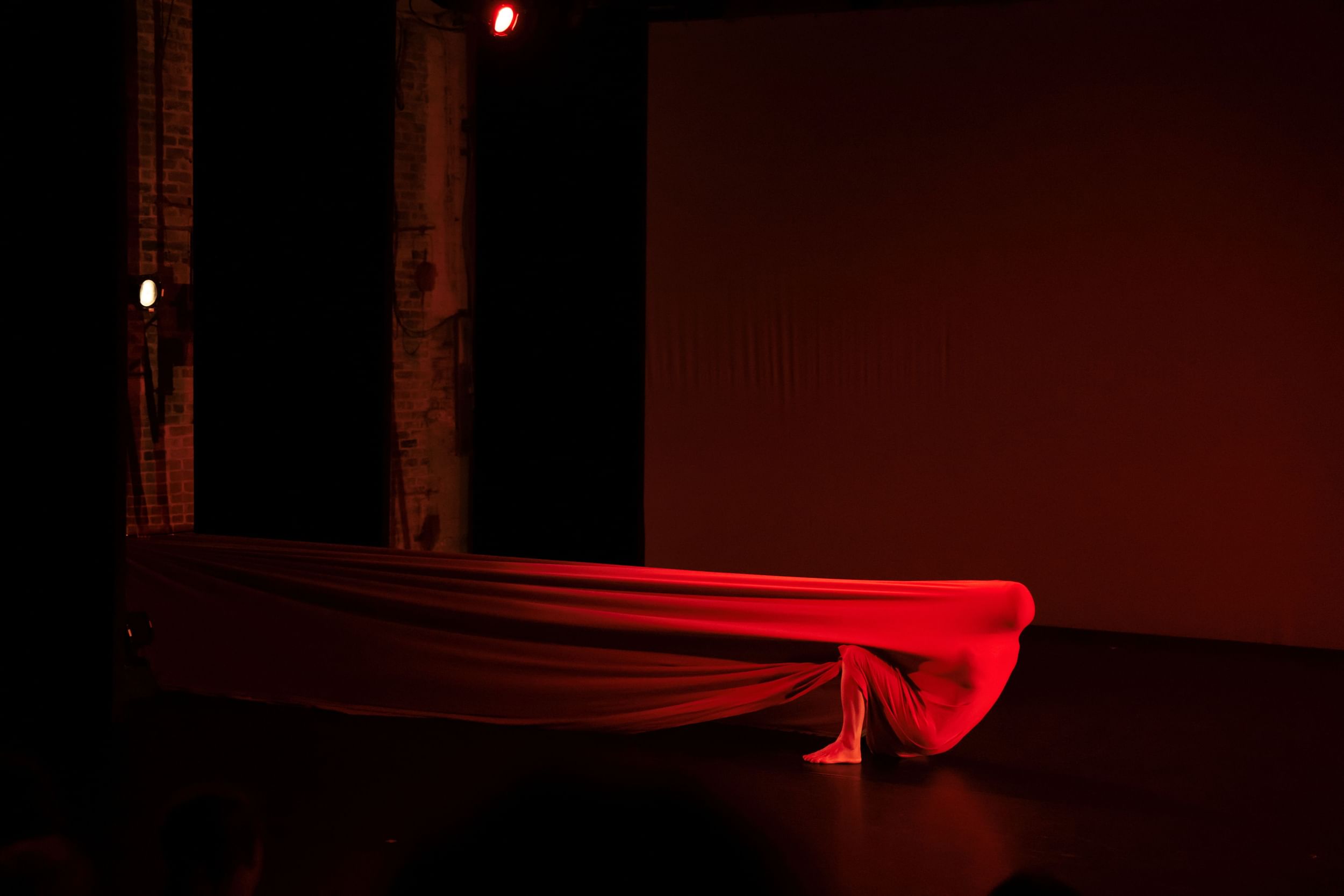 A red-lit dancer covered in a long piece of stretchy fabric anchored backstage pulls the fabric as they move on the floor