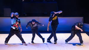 Four dancers in different poses on stage, dressed in black pants with print T-shirts.