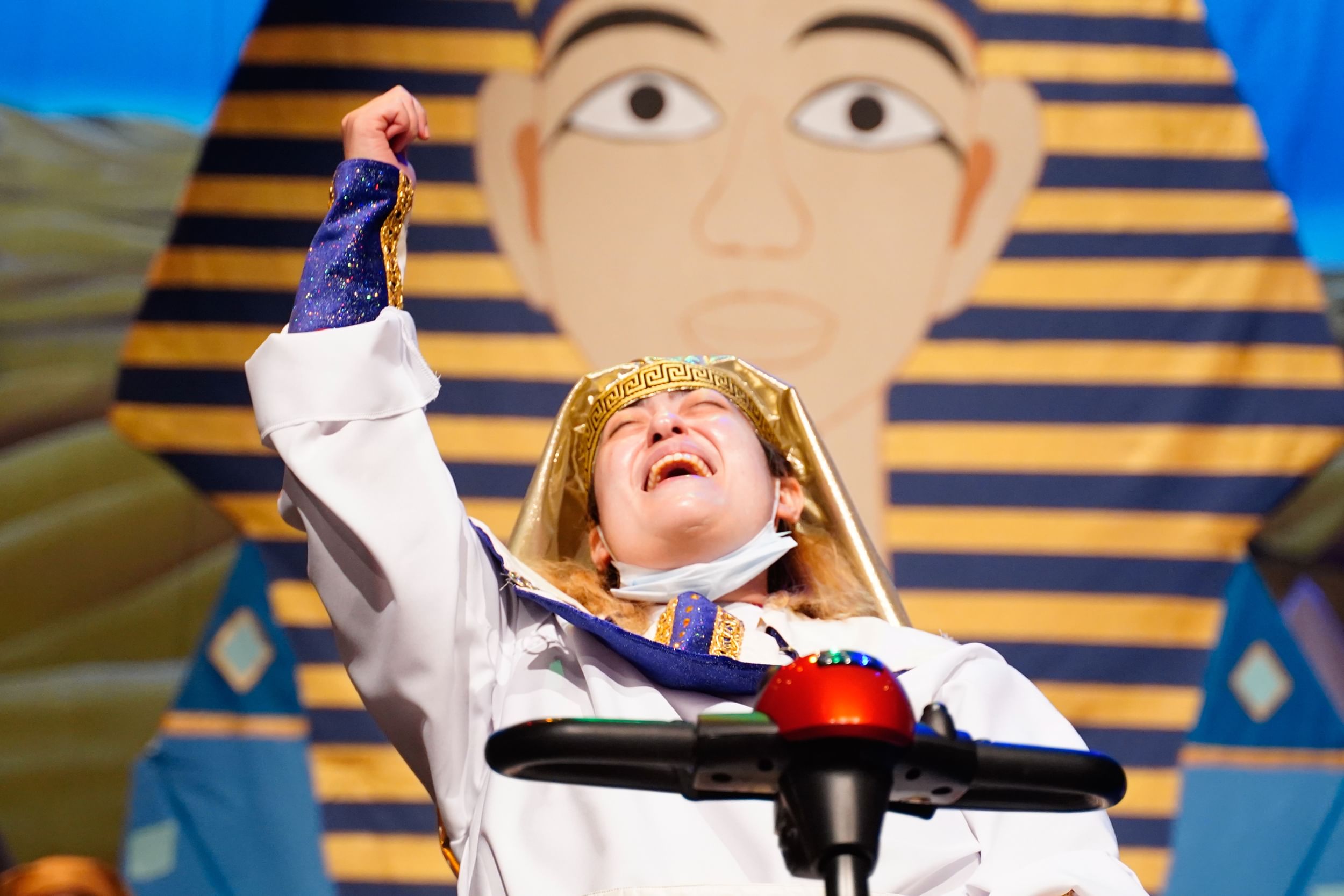 Closeup on Linden, in a white robe and golden headdress, tipping his head back ecstatically as he sings, one fist raised.