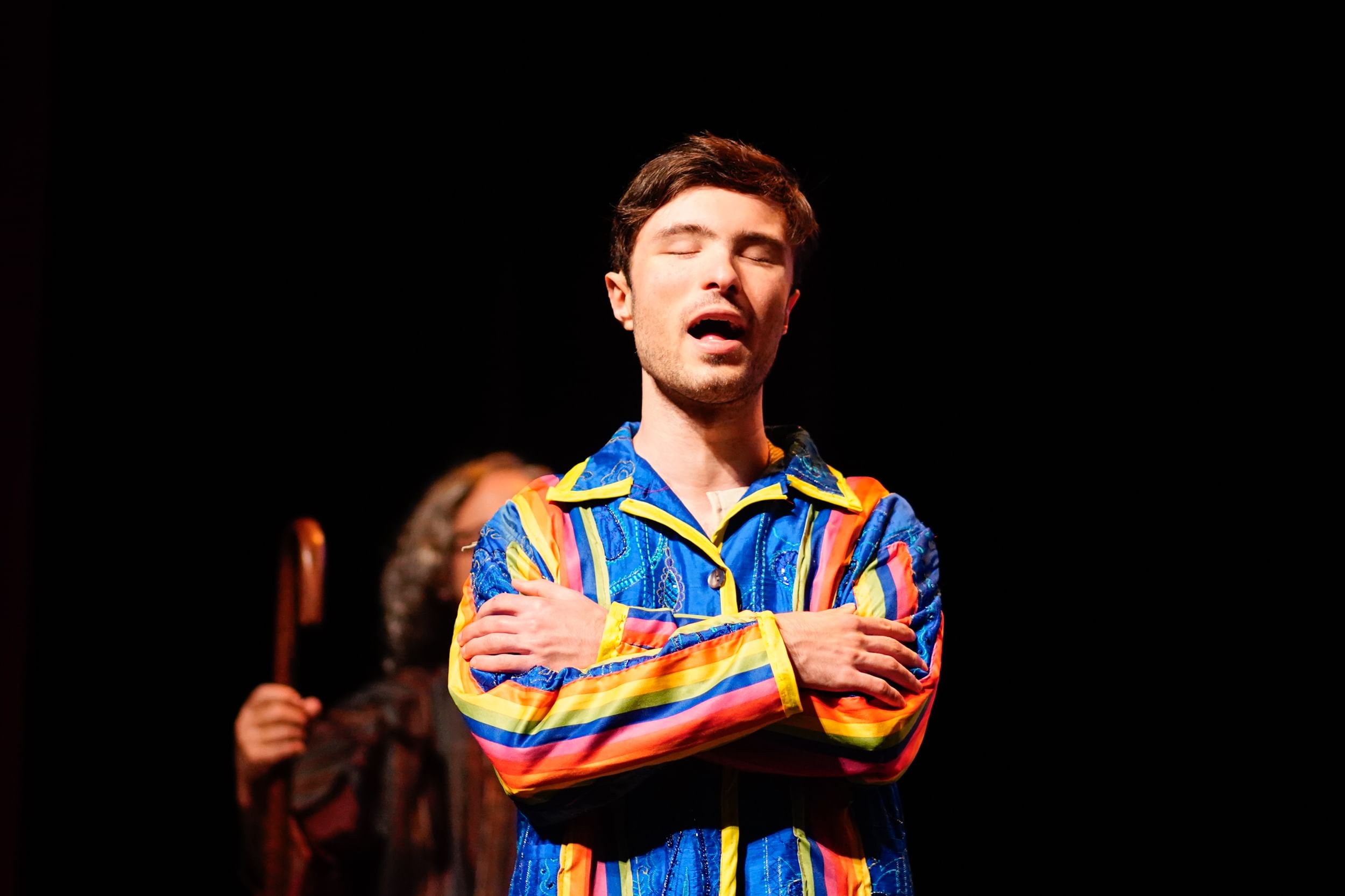 Bonenfant, a young white man with short brown hair wearing a colorful jacket, sings onstage with his arms crossed.