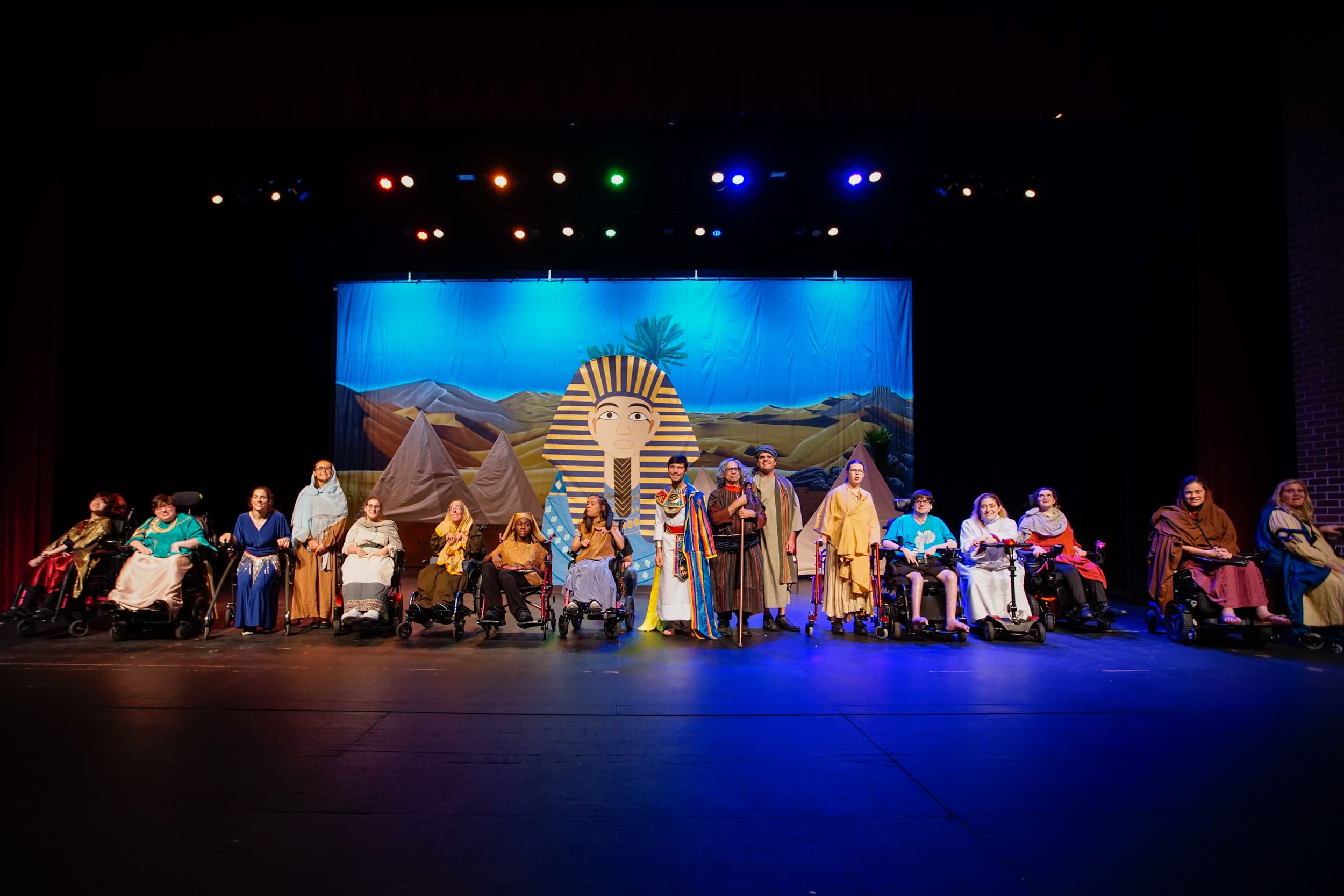 Costumed actors smile in a line onstage, many using wheelchairs, walkers, canes & scooters. A backdrop evokes ancient Egypt