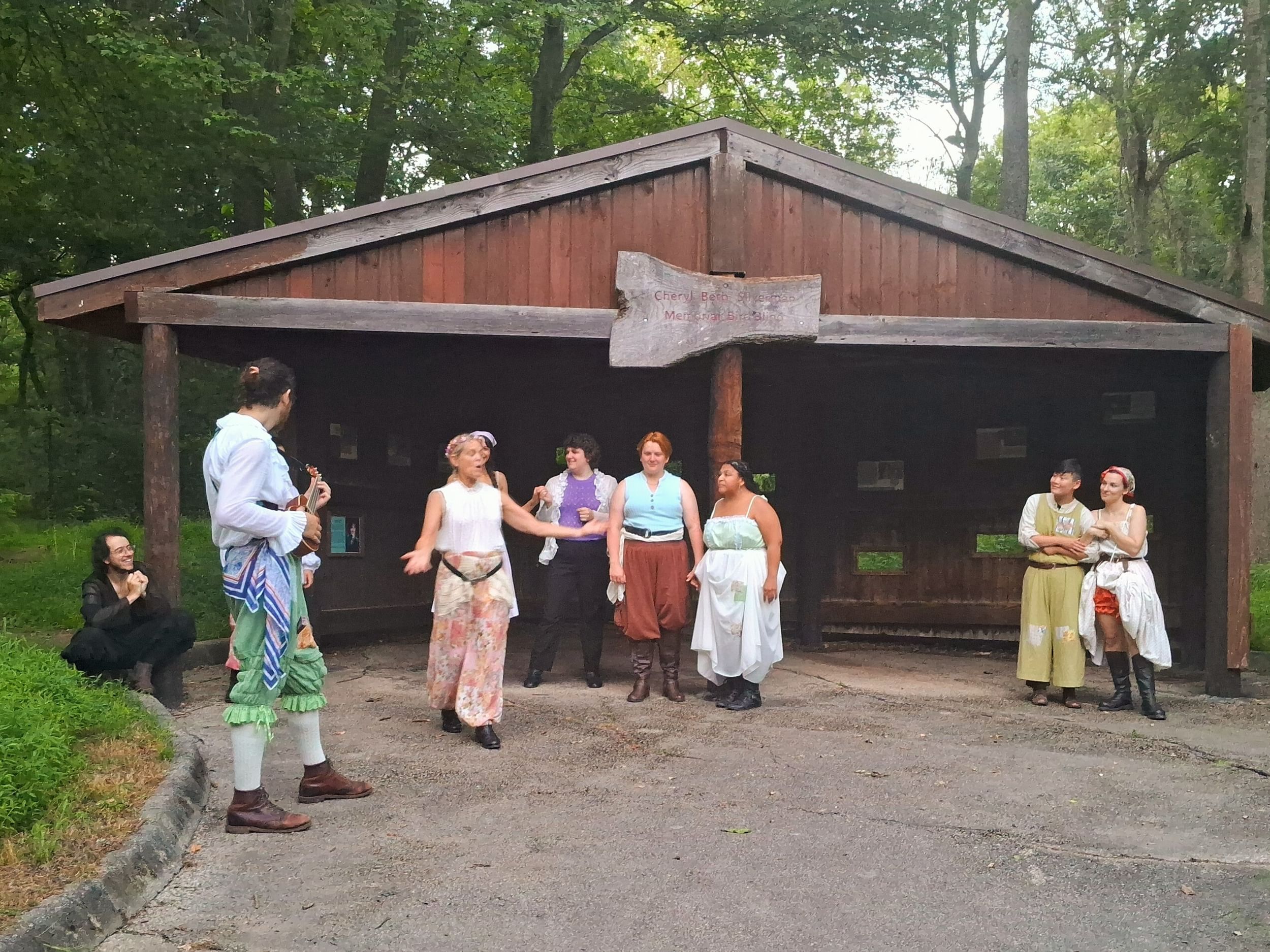Eight diverse actors in motley, flowing summer clothes perform outdoors in front of a large, open-fronted wooden shed.