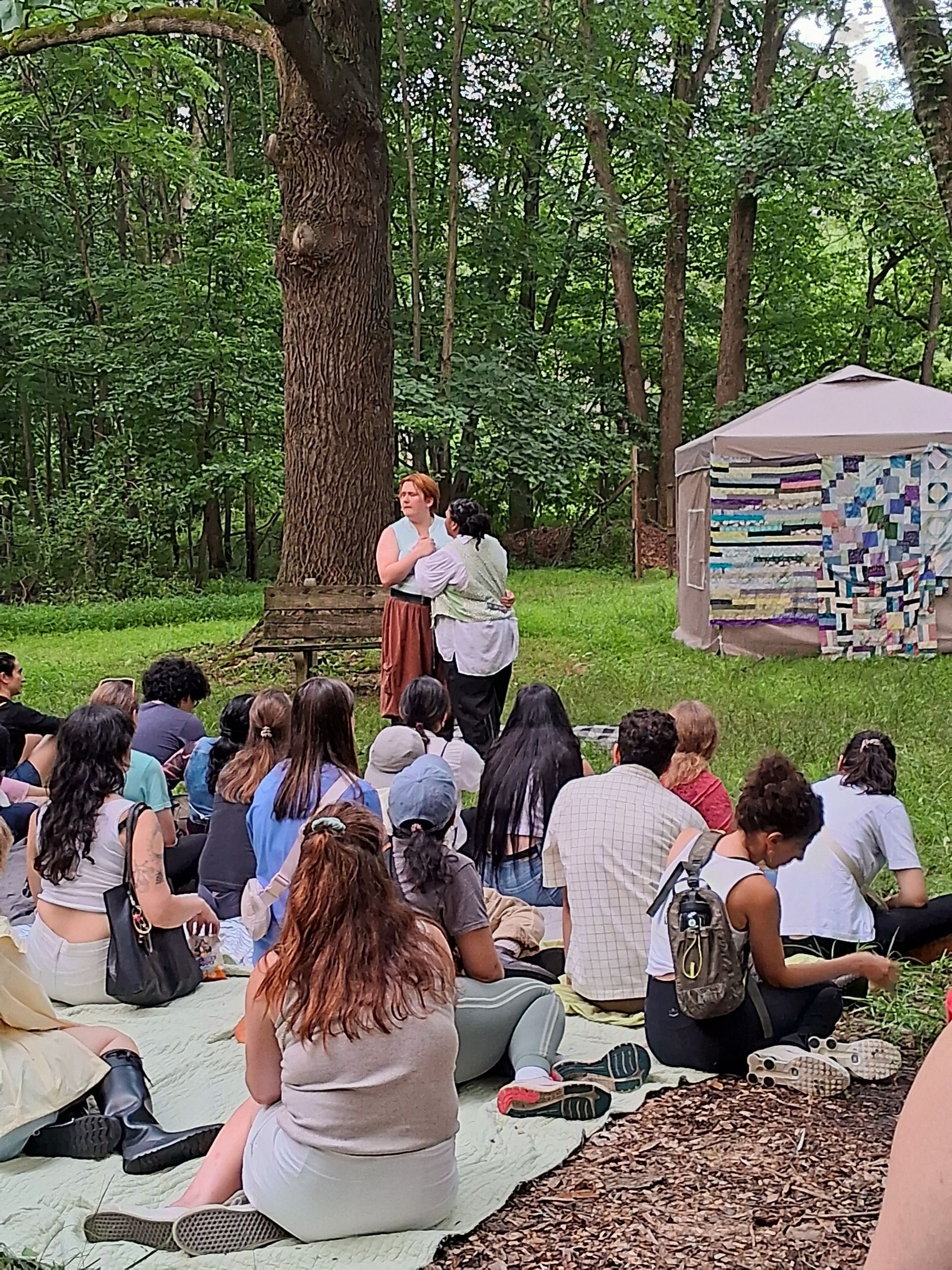 In a summer forest clearing, two actors perform in front of a few dozen people seated on blankets in front of them.