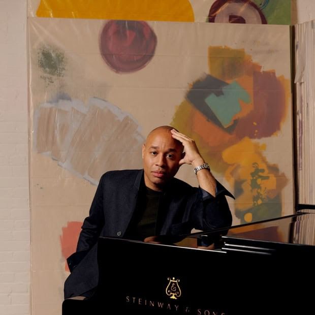 Diehl in black formalwear leans on one hand, elbow on the grand piano, a canvas with paint strokes behind him