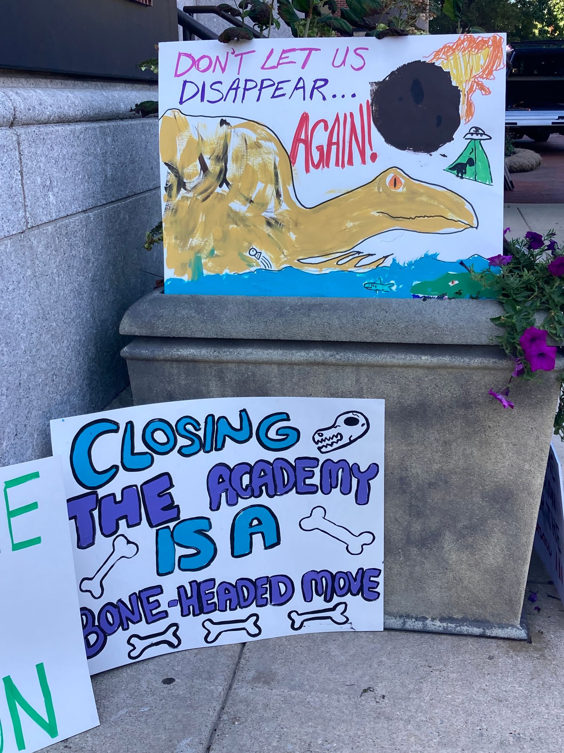 Two hand-drawn signs feature dinosaurs & bones, one reading “Closing the academy is a bone-headed move”