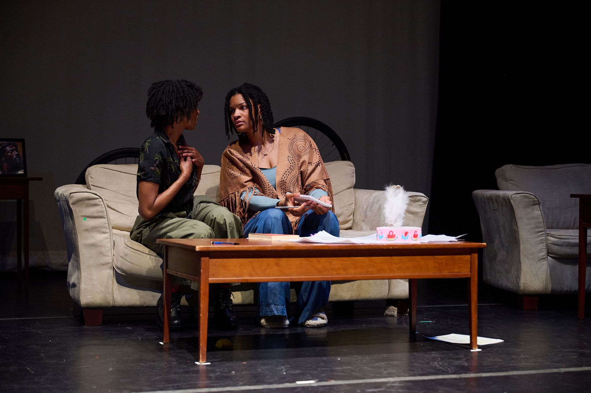 Charéce-Hall and Cynda Purnell, two Black women, sit together on a taupe couch on a black stage, talking seriously.