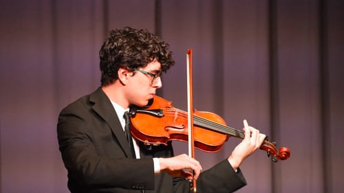 Close-up on Elbohy, a young man with dark curly hair and glasses, wearing a black suit, playing the violin.