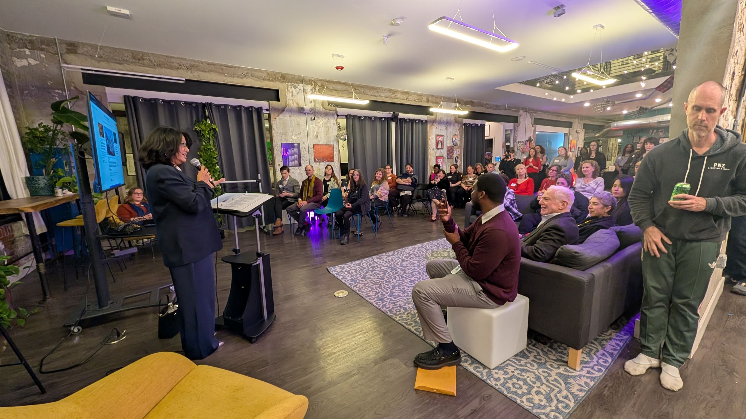 Ahmad, wearing a navy suit, speaks to a crowd of about 60 in a bright event space.