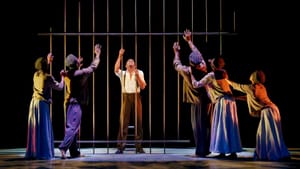 A male dancer at center, anguished behind bars. 4 others in patterned caps facing him, reaching on the other side of the bars