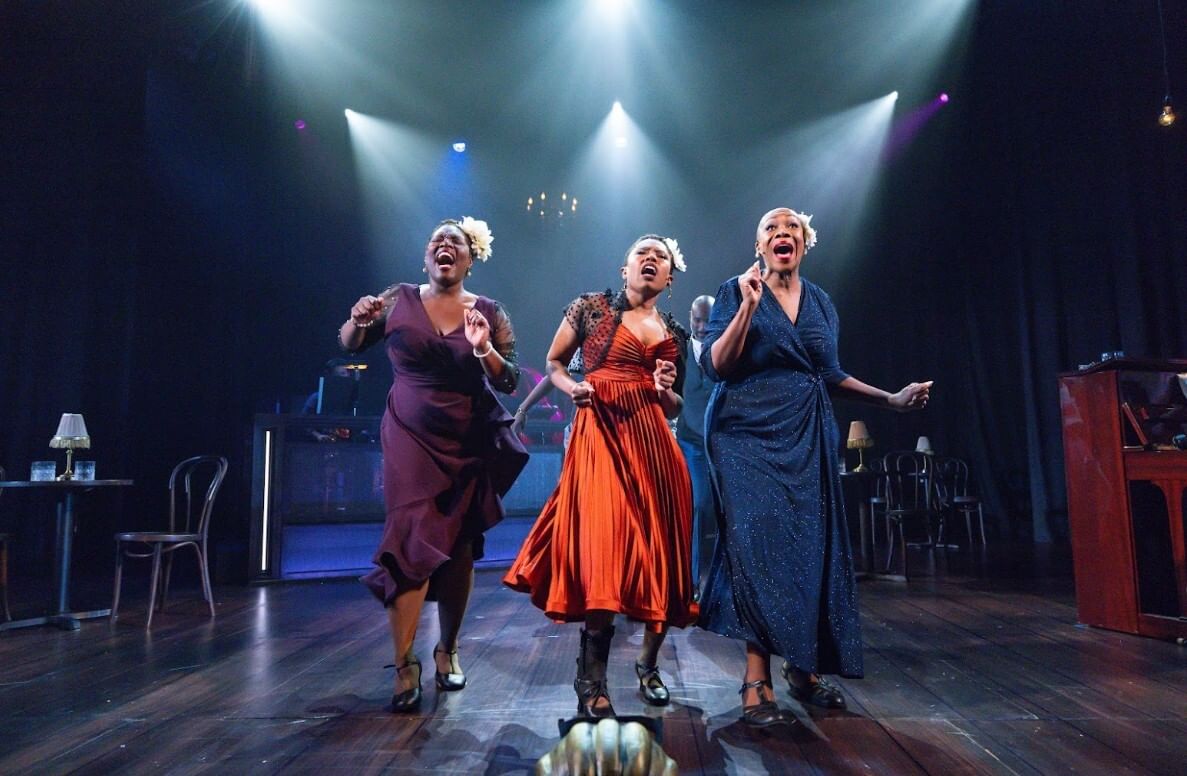 The three actors sing together at center, wearing richly draped evening dresses under a trio of spotlights.