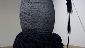 A giant textured gray ceramic vessel stands on a base of black acrylic hair, one hank of it attached to the gallery wall.