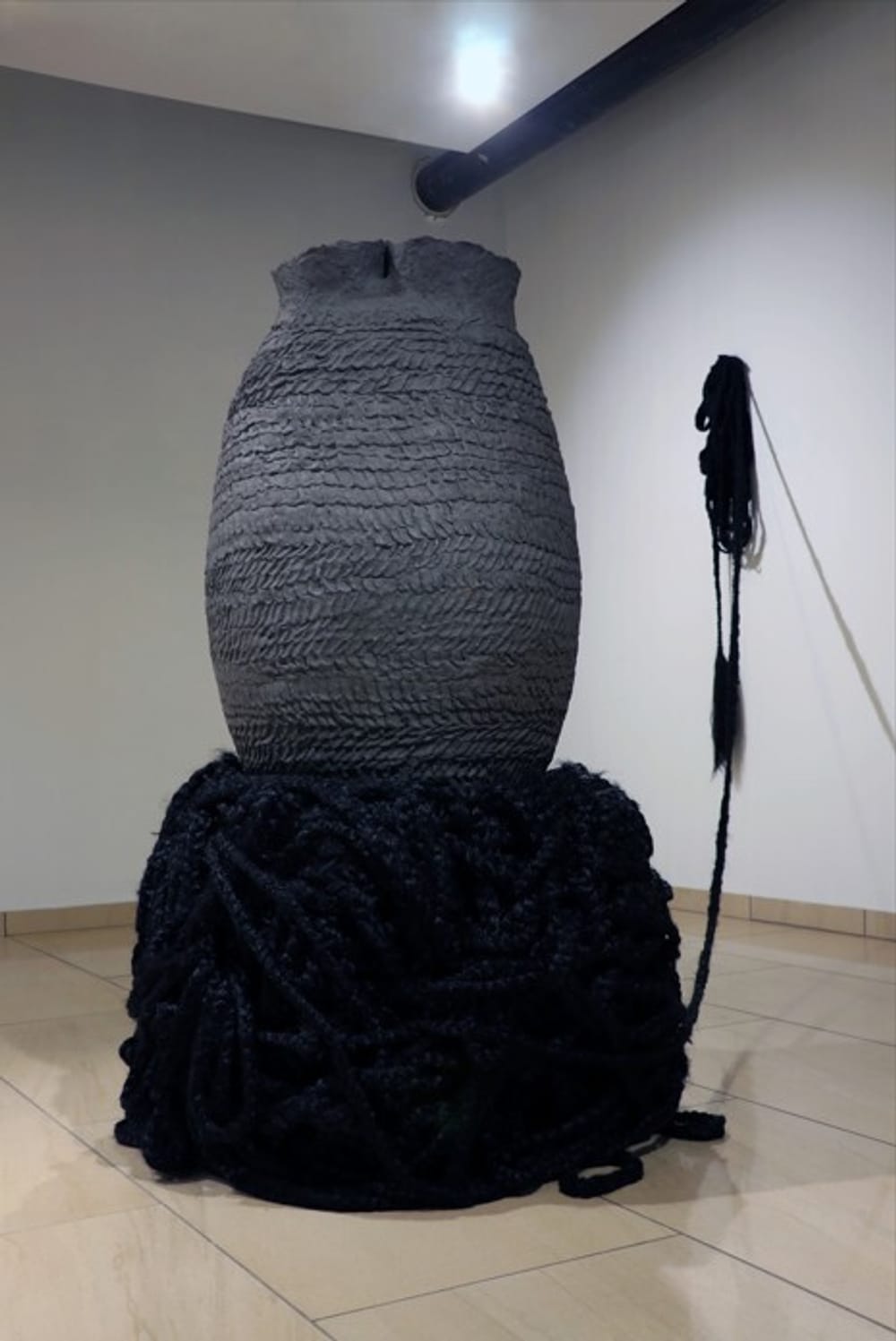 A giant textured gray ceramic vessel stands on a base of black acrylic hair, one hank of it attached to the gallery wall.