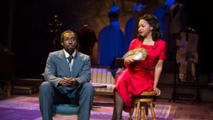 St. Clair, in a gray suit, and Dickinson, in a red dress, holding a fan, pose thoughtfully on low chairs onstage.