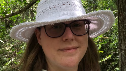 A selfie of Alaina, a 37-year-old white woman, in a green cypress swamp. She wears sunglasses and a touristy white hat.