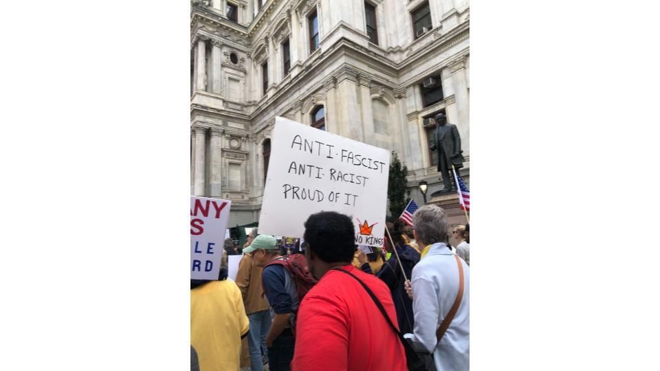 A Black protestor seen from behind holds a handwritten sign saying “Anti-fascist, anti-racist, proud of it” and “No Kings”