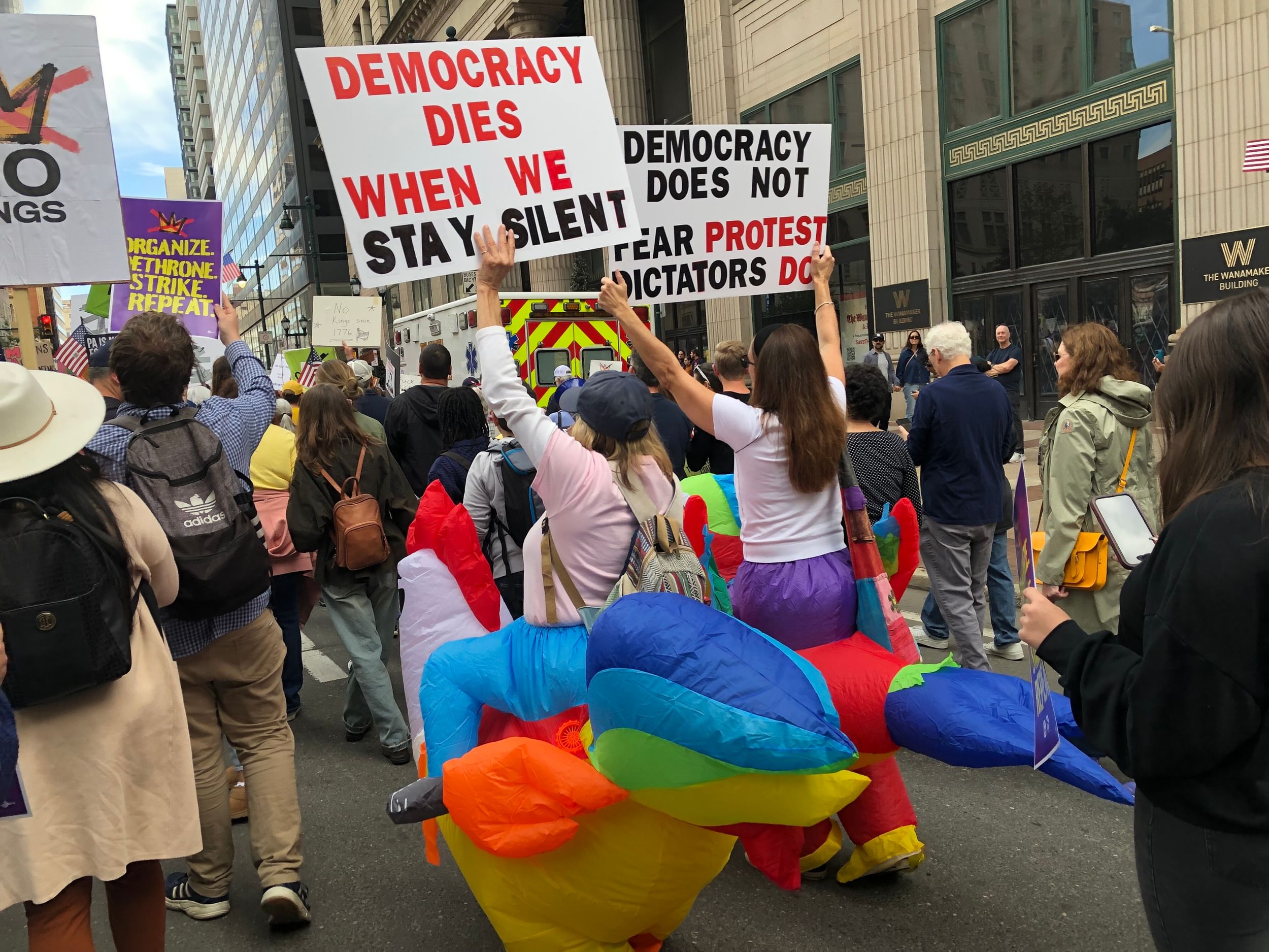 In a large protest crowd, two people in inflatable chicken suits hold large homemade signs with pro-democracy slogans.