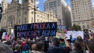 View of a huge protest crowd on a sunny day, centered on a giant sign that says MAKE AMERICA A DEMOCRACY AGAIN