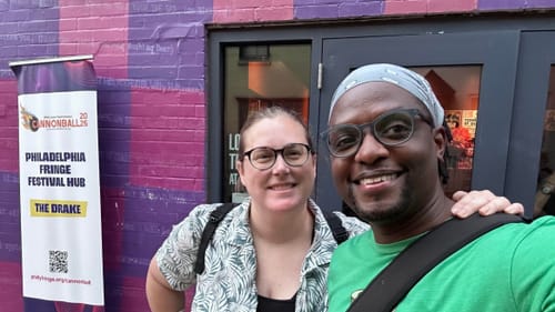 Alaina, a white woman, & Darnelle, a Black man, pose smiling together outside a theater decorated with a purple checkerboard