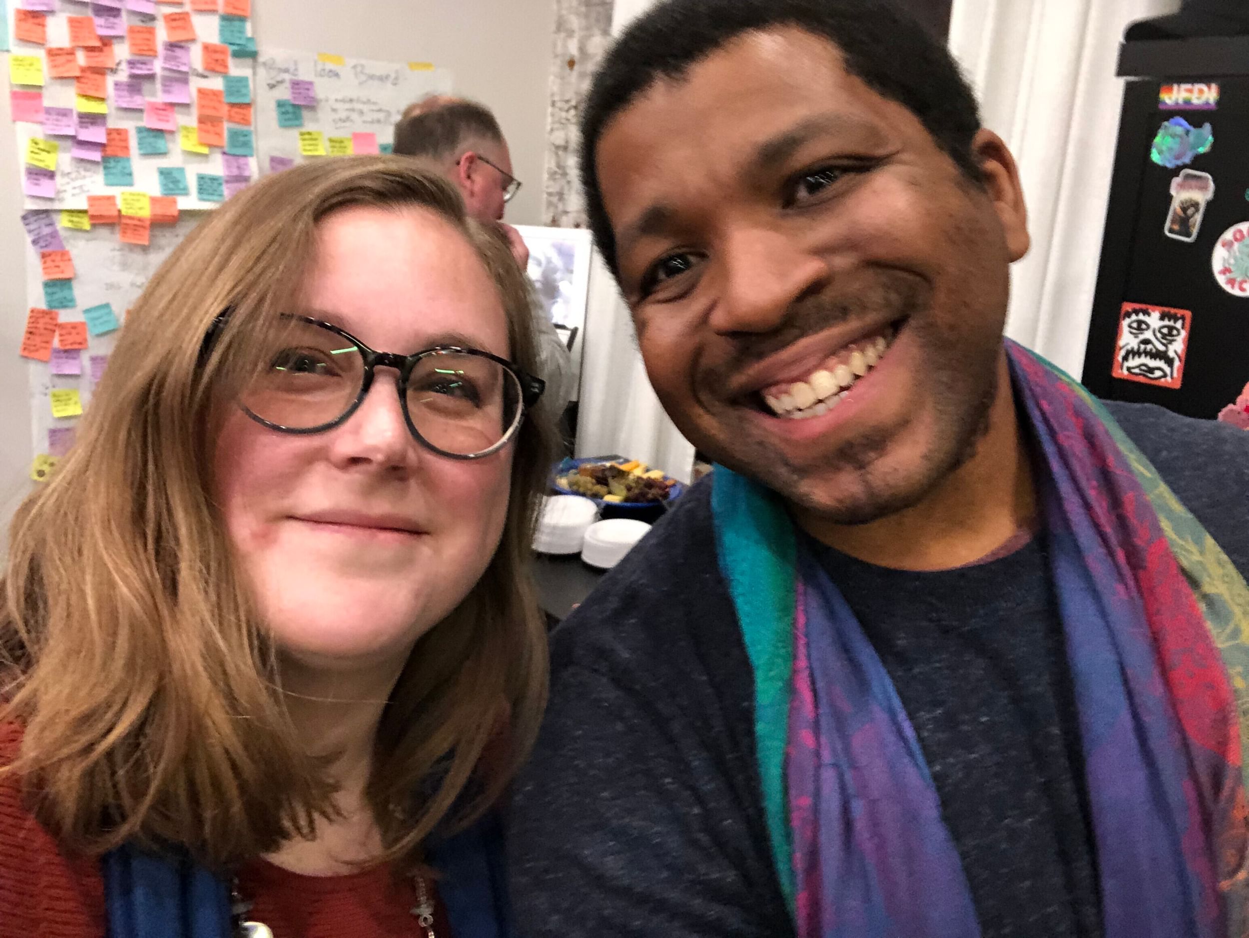 Alaina, a white woman with glasses and blond hair, smiles with Kyle, a Black man wearing a colorful scarf.