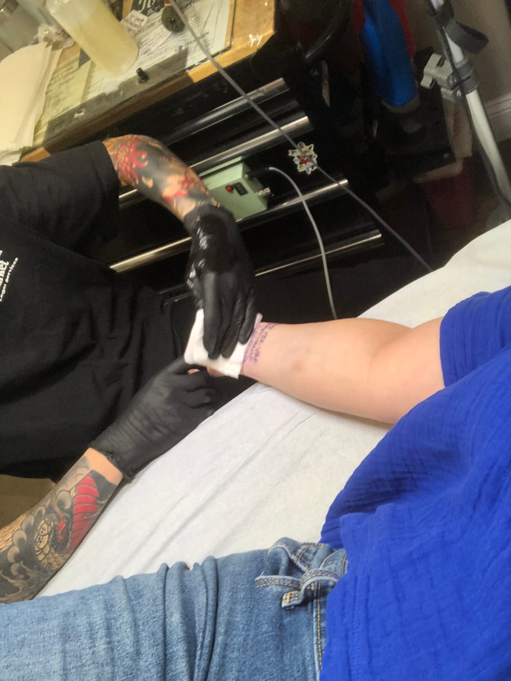 Alaina lies on the tattoo artist’s table while he finishes a tattoo on her right wrist. She’s wearing jeans and a blue shirt