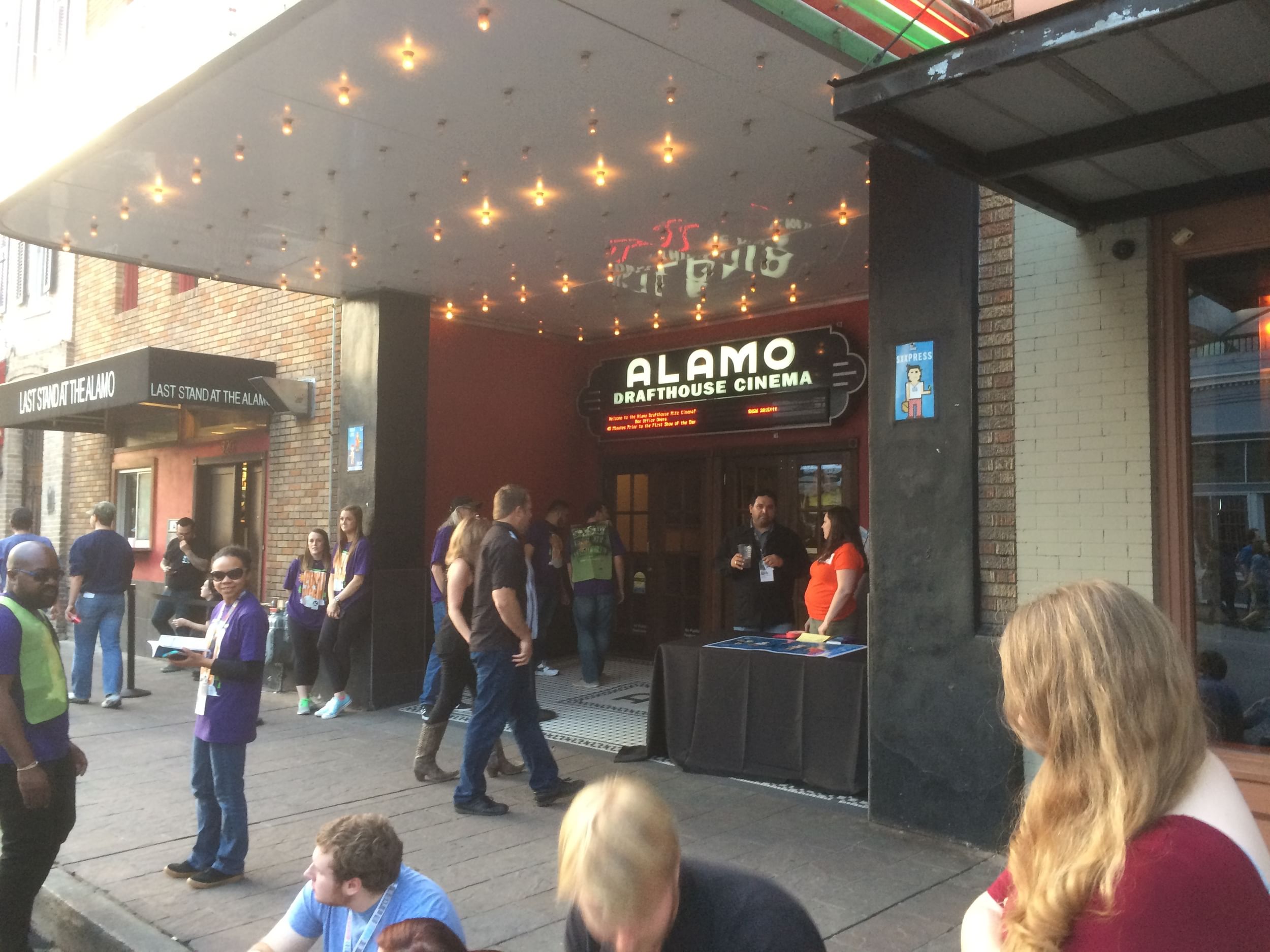 A small crowd of people move around outside the lit-up marquee of the Alamo Drafthouse entrance.
